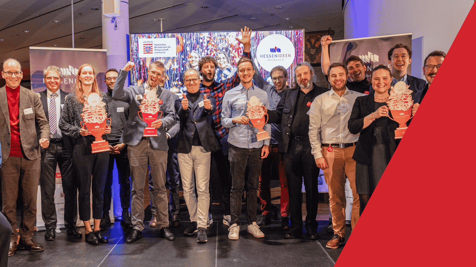 Auf dem Foto erkennt man 16 Personen. Sie stehen vor Hessen Ideen Bannern und jubeln in die Kamera. Eine Frau links, zwei Männer in der Mitte und eine Frau rechts halten jeweils einen Pokal hoch.